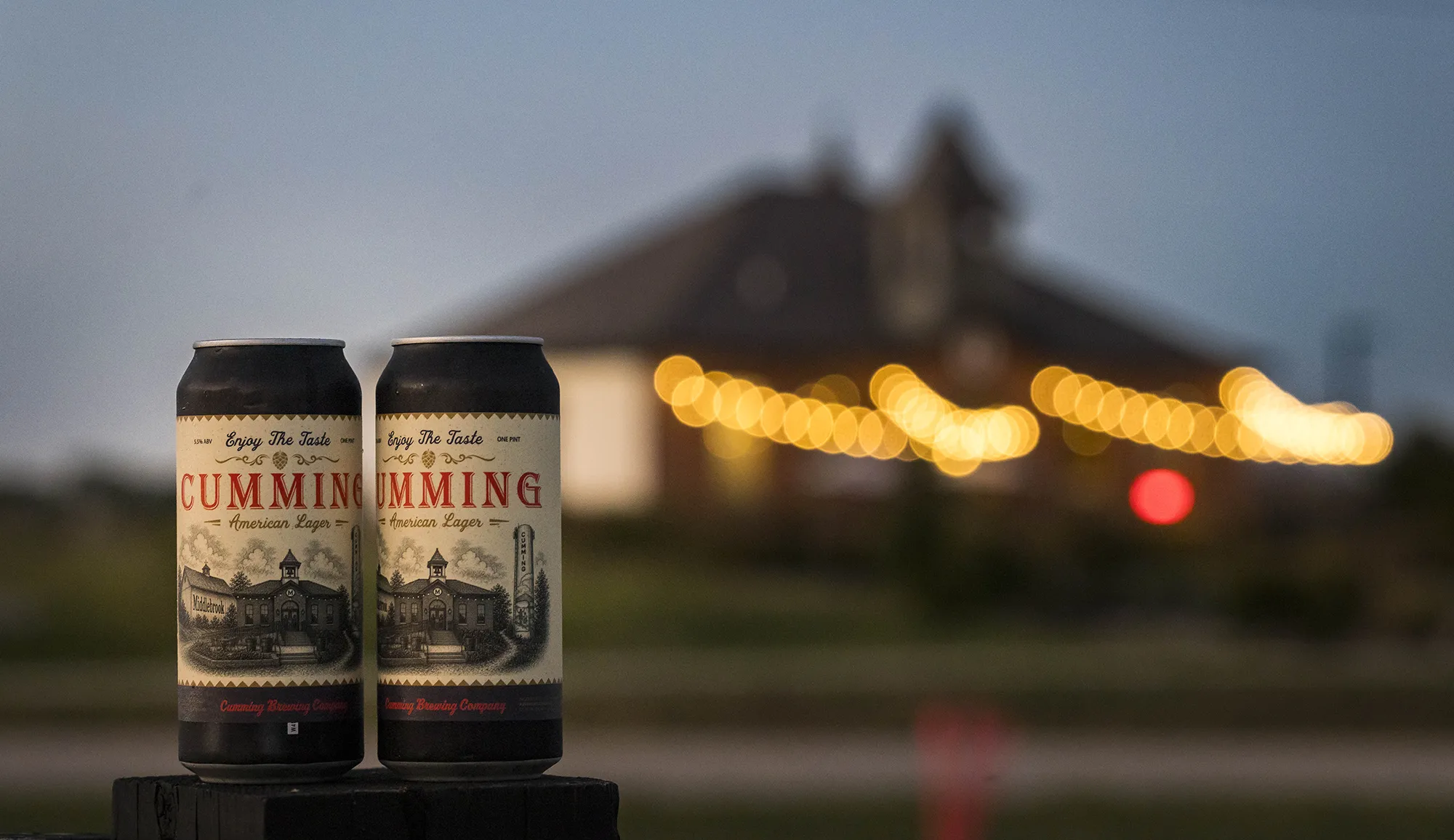 Two cans of Cumming American Lager at dusk with the Middlebrook Mercantile in the background