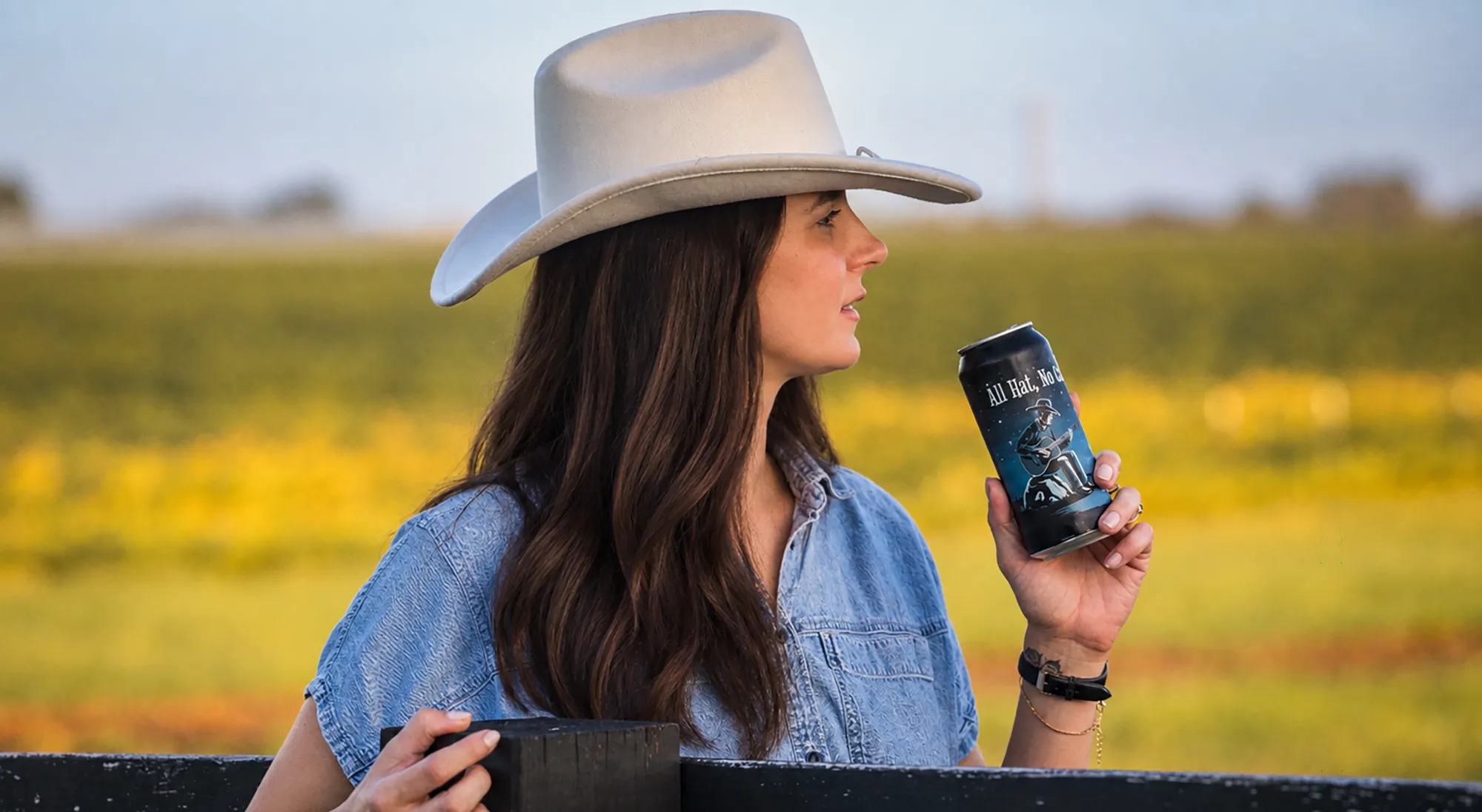 Woman in a cowboy hat holding a can of All Hat, No Cattle Coffee Stout in front of a field