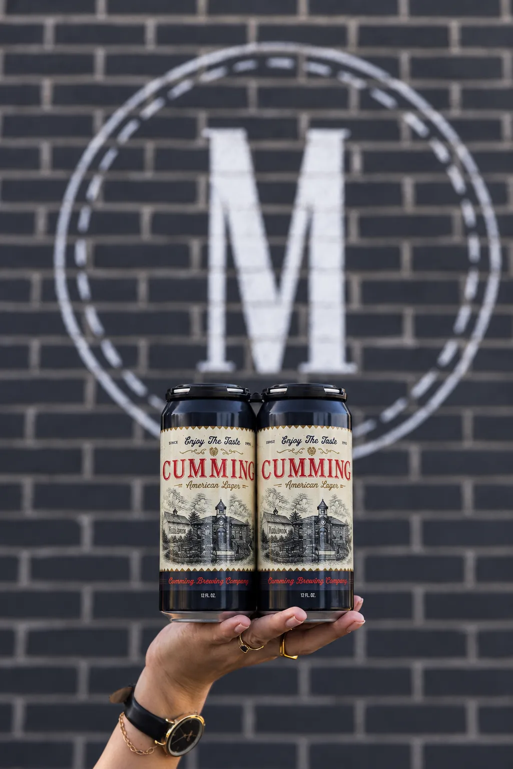 Hand holding two cans of Cumming American Lager in front of the Middlebrook M logo on a brick wall