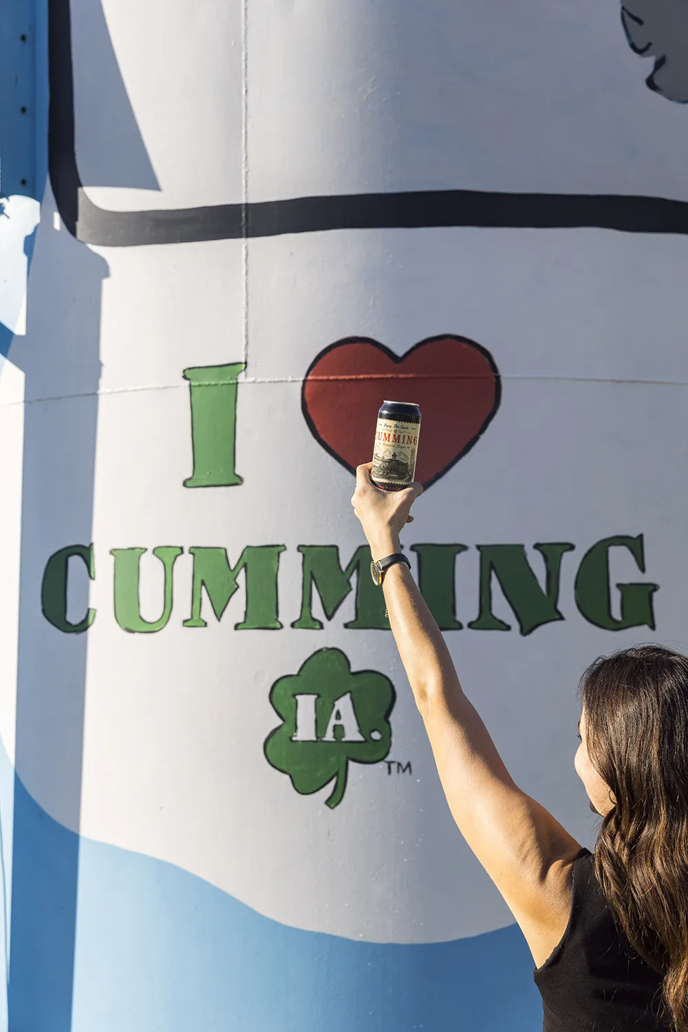 Woman holding a can of Cumming Lager up to the I Heart Cumming, Iowa mural on the water tower